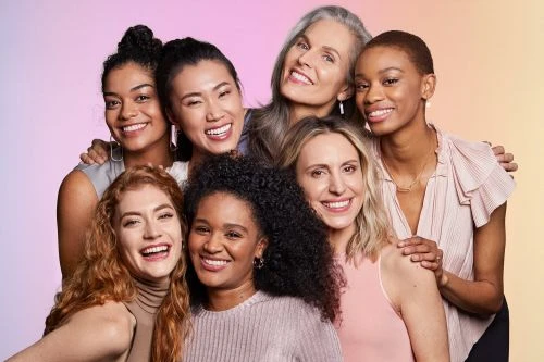 Group portrait of seven diverse women smiling and posing in front of a pastel gradient background in a beauty or skincare campaign.