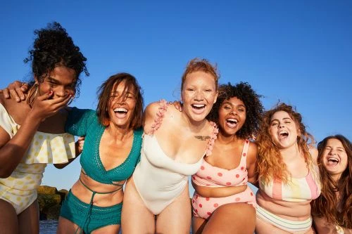 A group of six diverse women in swimwear joyfully embrace under a clear blue sky, radiating body positivity and summer vibes.