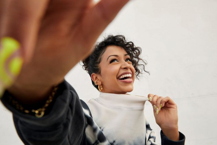 A smiling adult woman with curly hair wearing a tie-dye sweatshirt and gold jewelry reaches towards the camera in a brightly lit indoor setting.