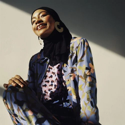 Smiling woman in hijab and floral outfit sits in sunlight, wearing statement earrings against a neutral background, fashion photography.