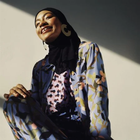 Smiling woman in hijab and floral outfit sits in sunlight, wearing statement earrings against a neutral background, fashion photography.