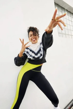 A woman in athletic apparel strikes a playful pose with hands outstretched, displaying a tie-dye sweatshirt and form-fitting leggings outdoors.