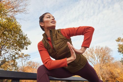 Woman in activewear stretches in park with fall foliage. She is on a bench, wearing a vest and leggings, balancing for fitness and flexibility.
