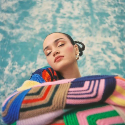 Portrait of a serene adult female with closed eyes, wearing a colourful knitted sweater against a blurred water background.