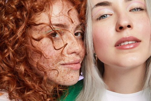 Close-up of two fair-skinned young adult women in a beauty context, featuring curly auburn hair and straight silver hair, plus makeup.