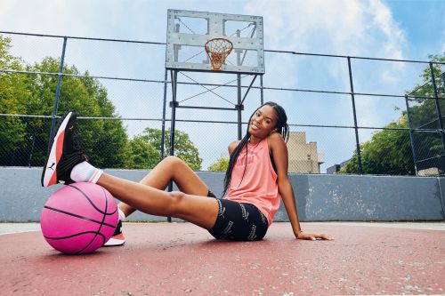 Woman in athletic wear with basketball on outdoor court near basketball hoop and trees, sitting against a blue sky.