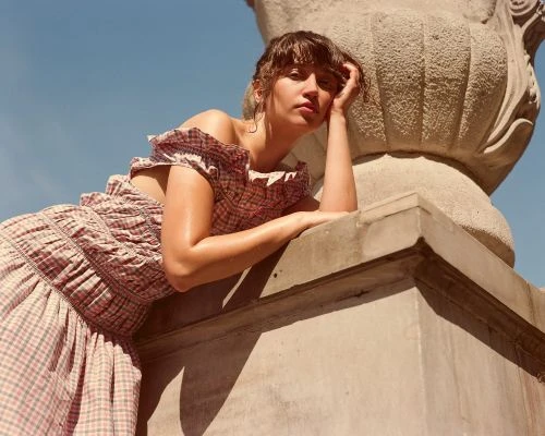 Woman in a pink gingham off-the-shoulder dress leaning against a stone structure under a clear blue sky
