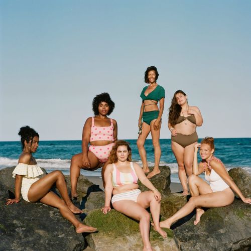 Diverse group of six women in vintage swimsuits pose on rocks at the beach with an ocean and blue sky background on a sunny day.