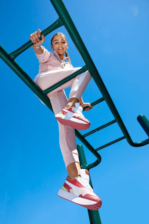 Young woman in pink tracksuit and red and white sneakers climbs green monkey bars against a clear blue sky, captured from a low angle.
