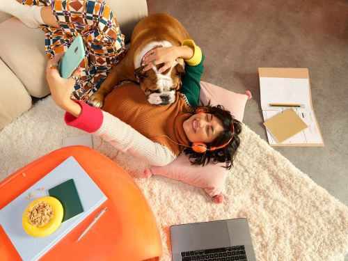 Woman lying on a rug taking a selfie with her phone, with her bulldog laying on her chest, next to a laptop, books, and a snack.