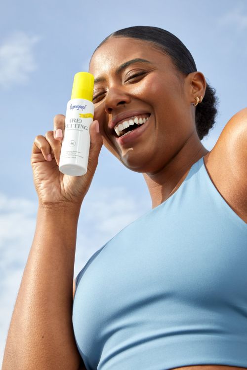 Smiling woman holds Supergoop! sunscreen setting spray in outdoor setting against bright sky, showcasing skincare and sun protection.