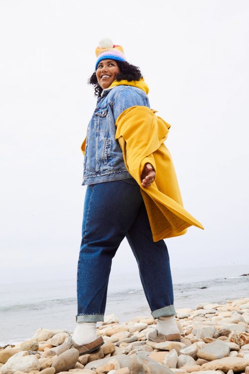 Adult woman in denim jacket, jeans, yellow coat, and beanie stands on a rocky beach against a light sky and water background.