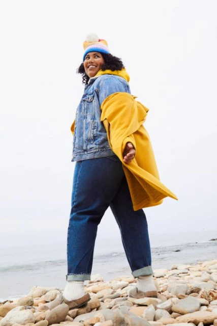 Adult woman in denim jacket, jeans, yellow coat, and beanie stands on a rocky beach against a light sky and water background.