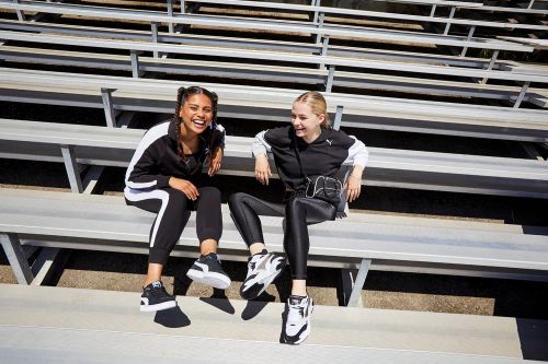 Two smiling women sit on stadium bleachers wearing matching black tracksuits and sneakers, enjoying an outdoor setting with natural light.
