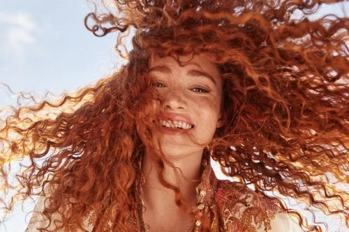 Close-up shot of a red-haired woman with freckles, smiling with windblown hair against a blue sky background on a bright, sunny day.