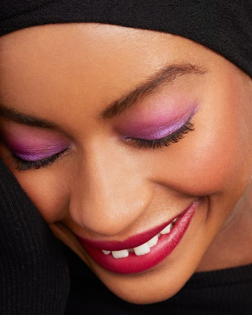 Close-up of a smiling African-American woman with closed eyes and purple eyeshadow, wearing a black head covering against a black backdrop.