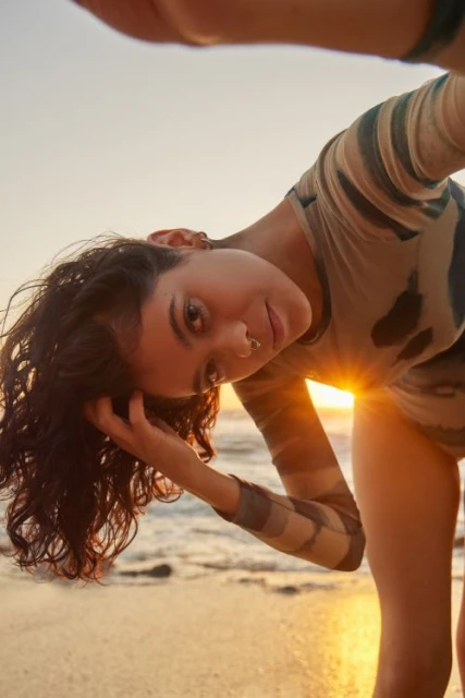 Young woman with curly hair poses at the beach during golden hour, wearing a patterned long-sleeve top, with ocean waves in background.