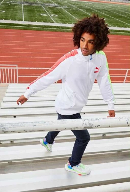 A man in a white Puma tracksuit and colorful sneakers walks up metal bleachers on an outdoor sports field with green turf.