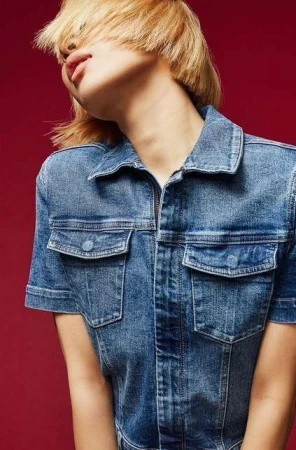 Model with blonde bob hairstyle wearing a blue denim jumpsuit against a solid dark red background