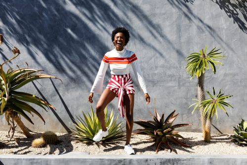 Woman in striped shorts and white top smiles outdoors with desert plants and a concrete wall casting shadows in bright sunlight.