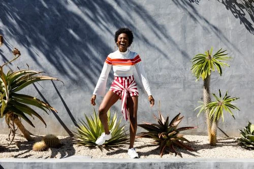 Woman in striped shorts and white top smiles outdoors with desert plants and a concrete wall casting shadows in bright sunlight.