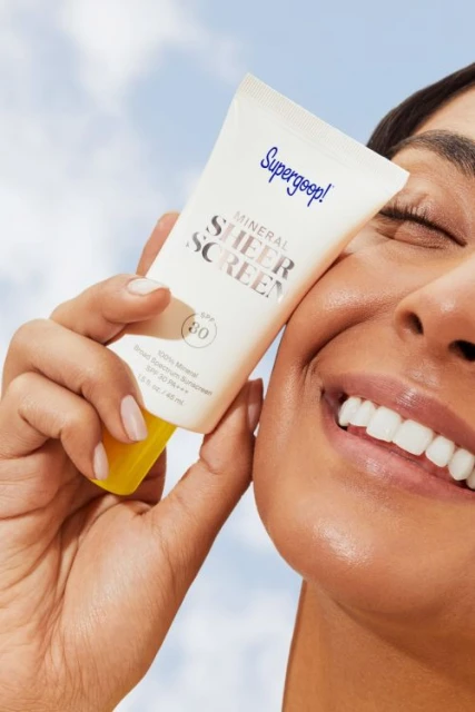 A woman smiles and holds a tube of Supergoop! Mineral Sheer Screen SPF 30 sunscreen, against a partly cloudy sky background.