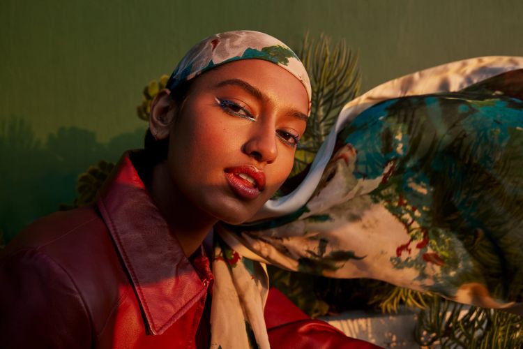 Close-up of a woman with dramatic eye makeup and a patterned headscarf, shot in warm light against a green and tan backdrop.
