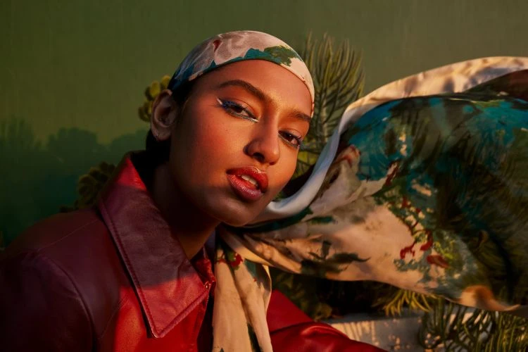 Close-up of a woman with dramatic eye makeup and a patterned headscarf, shot in warm light against a green and tan backdrop.