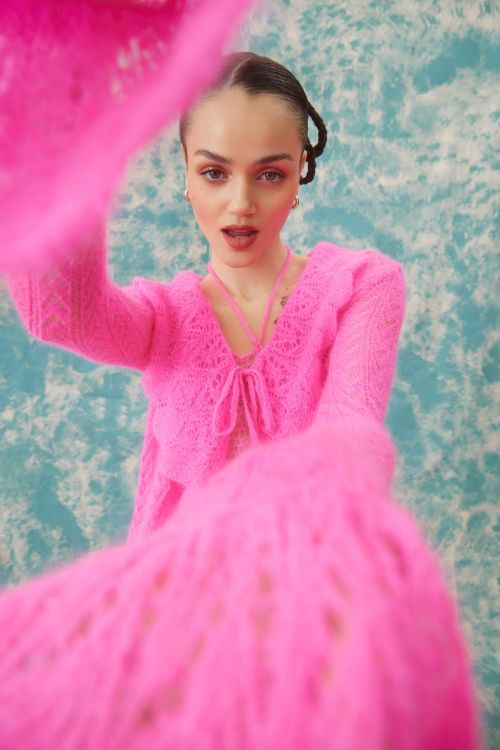 A young woman is photographed in a bright pink knit outfit against a cloudy light blue background in a fashion forward style.