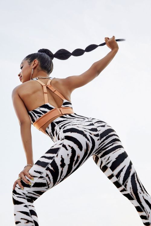 Adult woman with zebra print leggings and sports bra with tan straps and a high ponytail poses against a bright, clear sky.