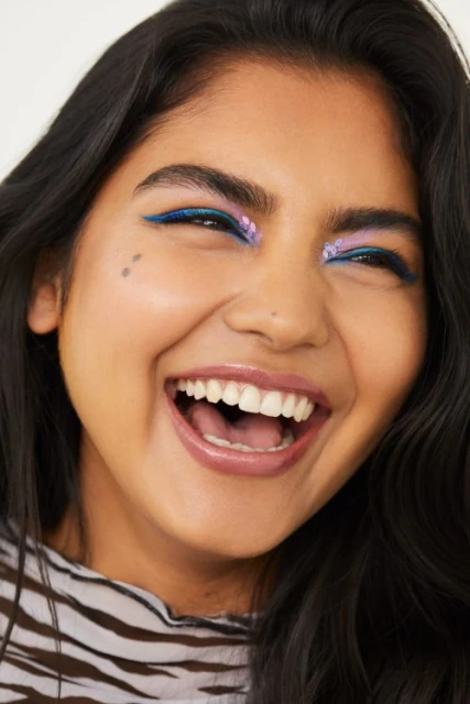 Close-up of a smiling woman with dark hair, wearing colorful makeup with a winged eyeliner and flower details for a beauty advertisement.