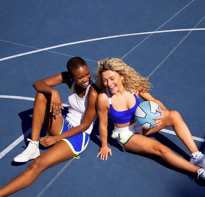 Two women smile sitting on a blue sports court. One holds a soccer ball. They wear coordinated athletic shorts and sports bras.