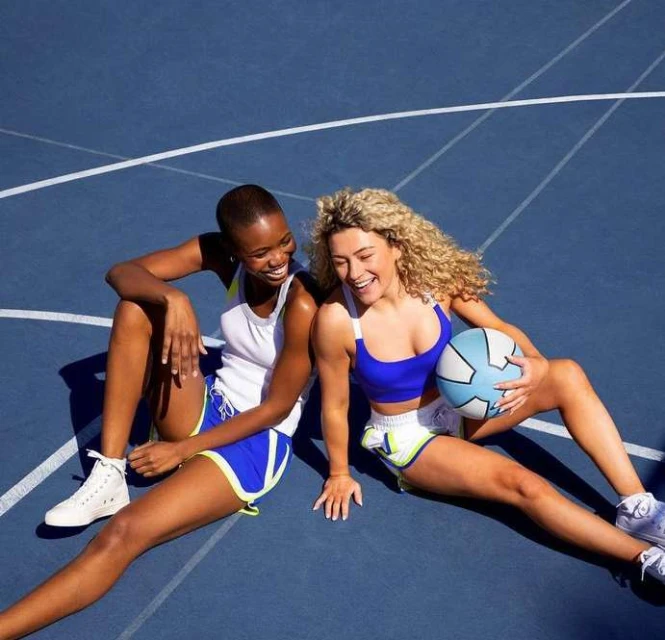 Two women smile sitting on a blue sports court. One holds a soccer ball. They wear coordinated athletic shorts and sports bras.