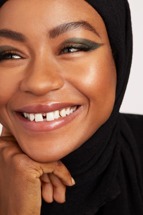 Close-up of a smiling woman wearing a black headscarf, showcasing olive-green eyeliner and radiant skin in a beauty-focused image.