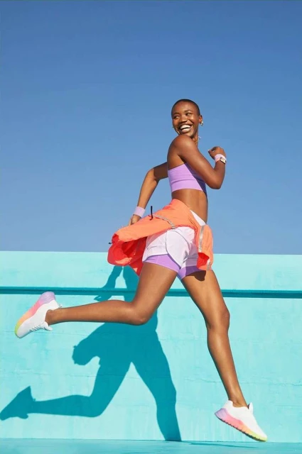 Smiling woman in athletic wear mid-stride against a blue background, wearing a lilac sports bra, white shorts, and an orange jacket.