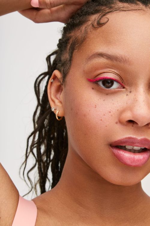 Close-up portrait of a young woman with braided hair and pink eyeliner, posing against a white background in a beauty context.