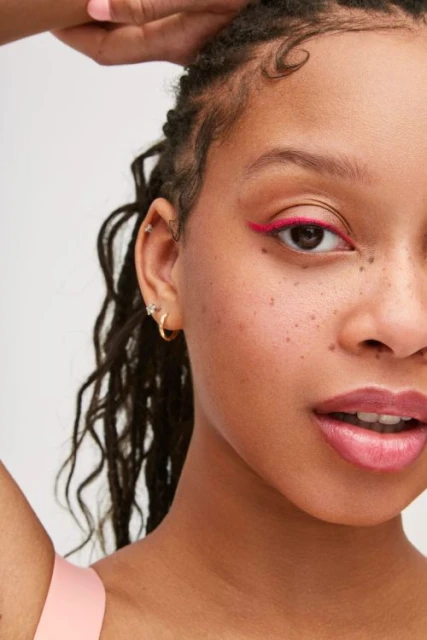 Close-up portrait of a young woman with braided hair and pink eyeliner, posing against a white background in a beauty context.