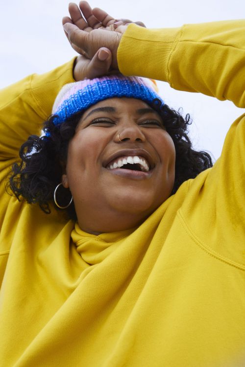Close-up of a smiling woman with curly hair, wearing a colorful knit hat and a bright yellow sweatshirt, posing indoors.