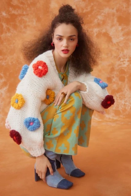 Woman with curly hair posing in a colorful patterned outfit with a flower-embellished sweater in front of a mottled orange backdrop.