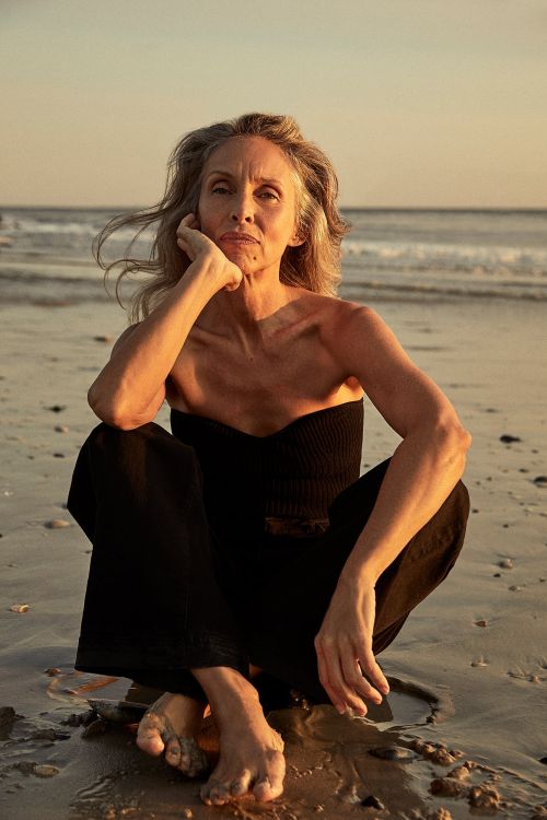 Older woman with gray hair in black outfit sits cross-legged on sandy beach with the ocean and sky in the background at sunset.
