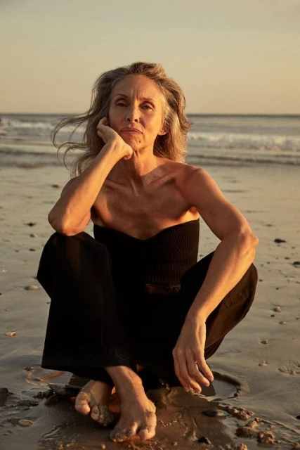 Older woman with gray hair in black outfit sits cross-legged on sandy beach with the ocean and sky in the background at sunset.