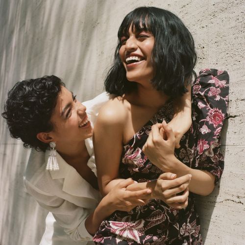 Two smiling women embrace in an outdoor fashion setting. One woman wears a floral dress, the other a white blazer, against a textured wall.