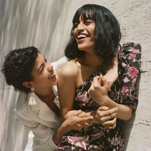 Two smiling women embrace in an outdoor fashion setting. One woman wears a floral dress, the other a white blazer, against a textured wall.