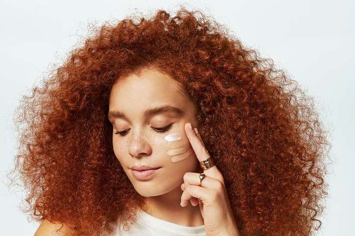 Woman with red curly hair applies different shades of foundation to her face, testing the colors. Beauty shot in front of a light backdrop.
