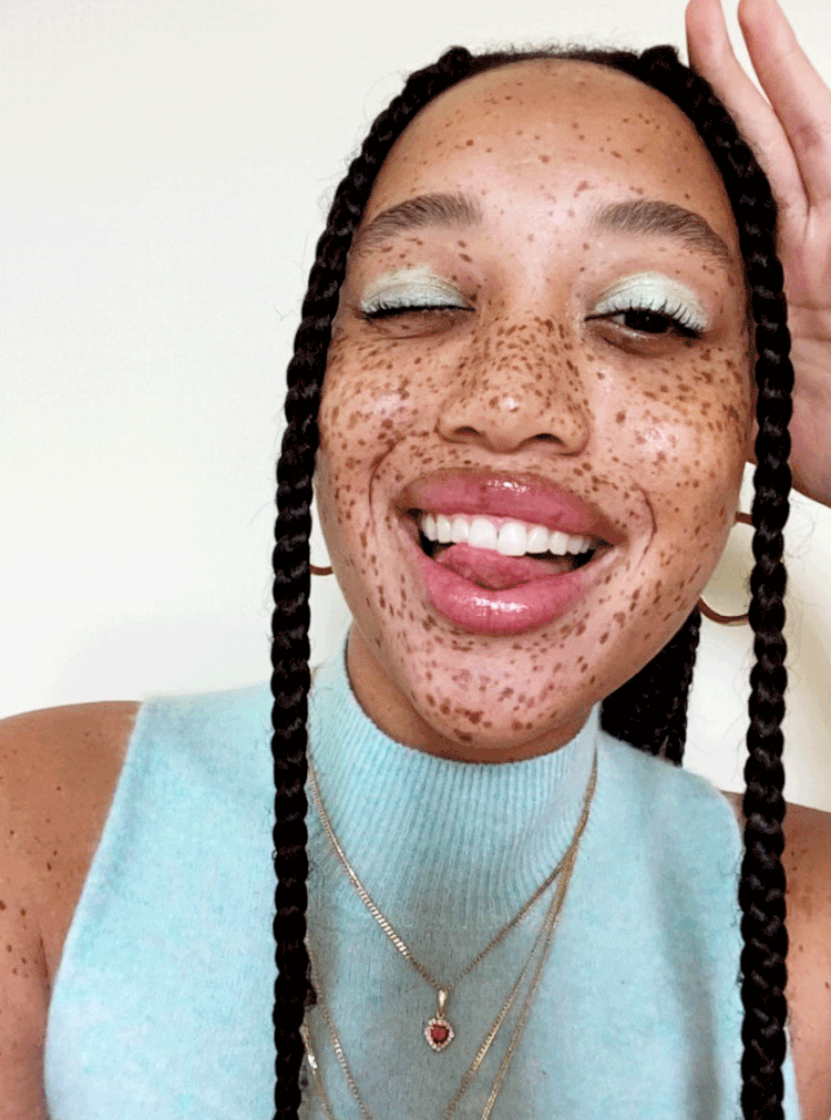Close-up of an adult woman with freckles and braids wearing light blue top, gold hoop earrings, and necklaces, smiling and sticking out tongue.