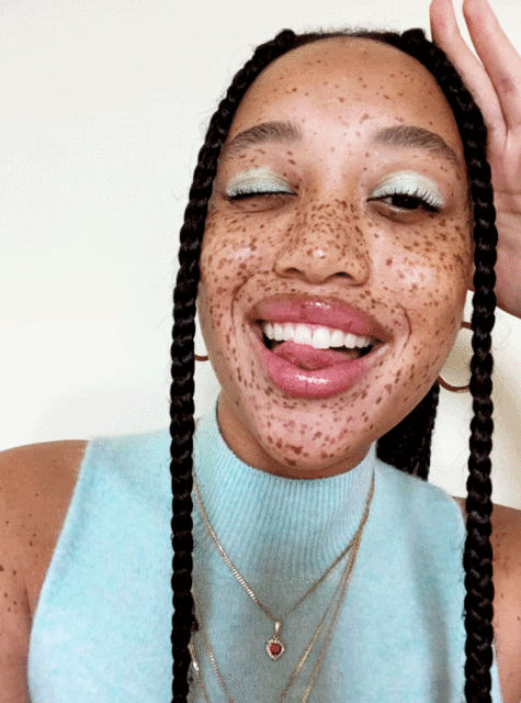 Close-up of an adult woman with freckles and braids wearing light blue top, gold hoop earrings, and necklaces, smiling and sticking out tongue.