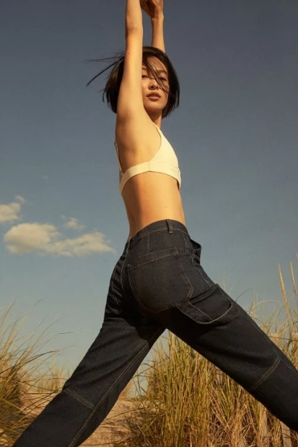 Woman in dark denim carpenter pants and a light-colored crop top stands outside against a blue sky and tall grass.
