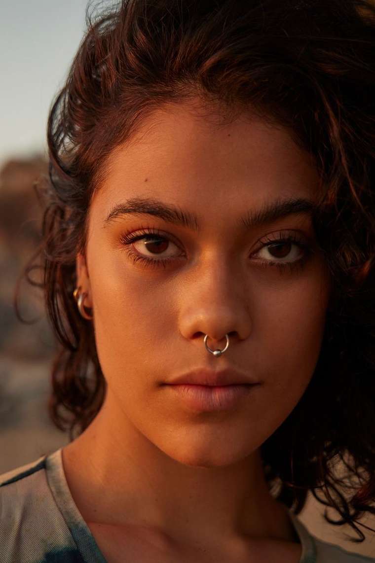 Close-up portrait of an adult woman with curly hair and a nose ring, warm lighting, against a blurry outdoor background at sunset.