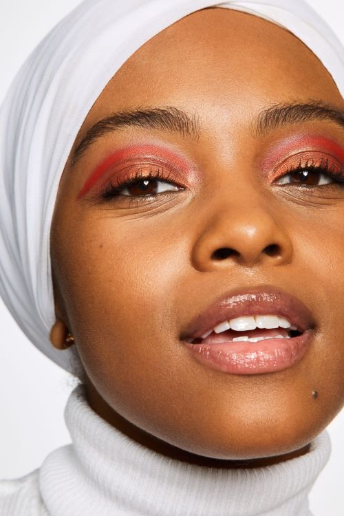 Close-up portrait of a woman with dark skin and red eyeshadow wearing a white hijab and turtleneck sweater in a studio setting.