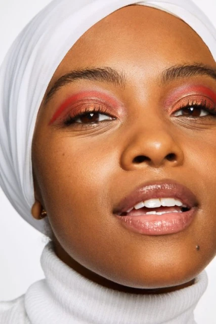 Close-up portrait of a woman with dark skin and red eyeshadow wearing a white hijab and turtleneck sweater in a studio setting.